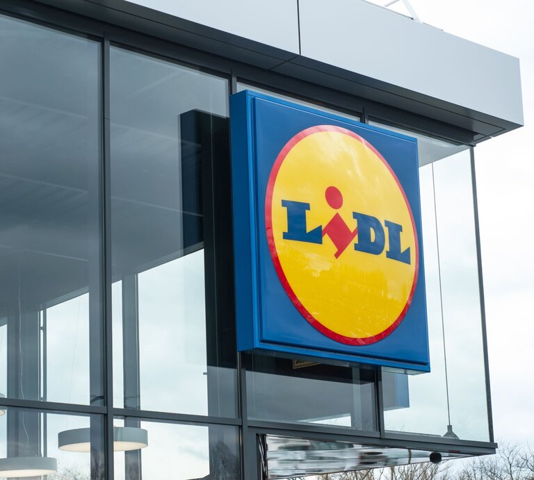  Das Bild zeigt das typische Logo des Einzelhändlers Lidl an einem Geschäftsgebäude. Die moderne Architektur des Gebäudes mit großen Glasflächen und der klare Himmel im Hintergrund vermitteln einen zeitgenössischen Eindruck. Lidl ist bekannt für sein Discount-Supermarktkonzept, das in vielen Ländern vertreten ist und eine breite Palette von Produkten, darunter Lebensmittel, Haushaltswaren und saisonale Artikel, zu günstigen Preisen anbietet. Das präsentierte Logo mit dem blauen Hintergrund und dem gelben Kreis mit roter Schrift ist sofort erkennbar und repräsentiert die Marke Lidl weltweit. | © Adobe Stock/ pixarno
