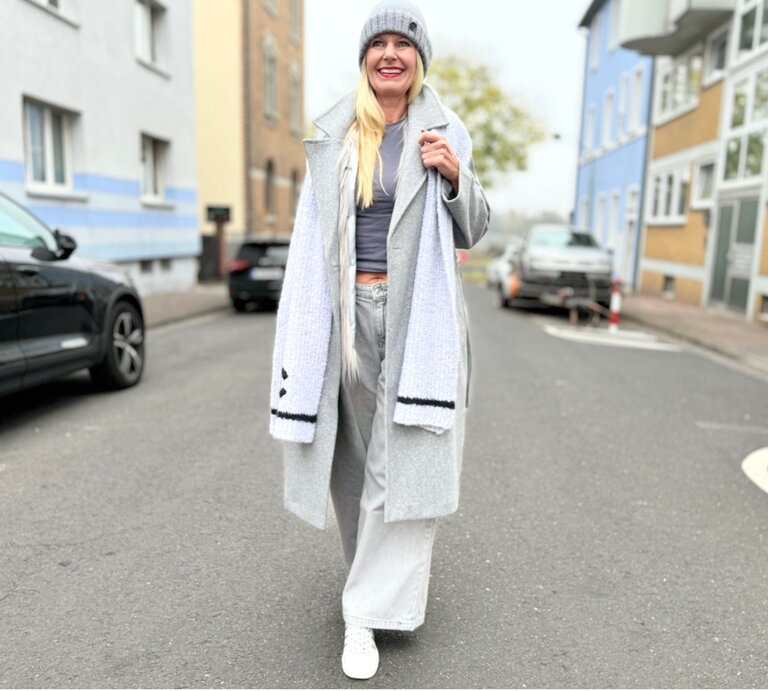 Lächelnde Frau mit grauem Mantel, weiter Hose und Mütze läuft an einem bewölkten Tag durch eine Stadtstraße mit bunten Fassaden. | © Renate Zott