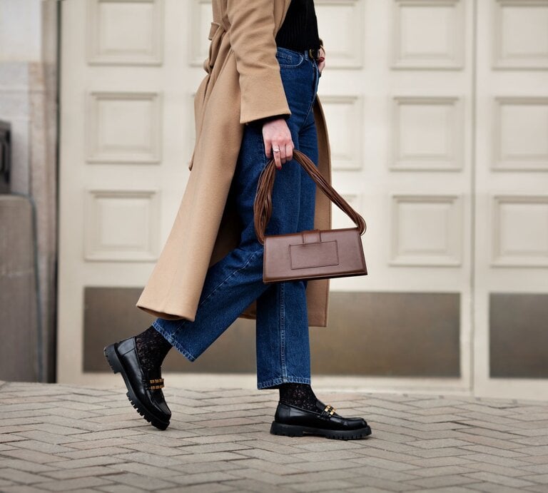 Frau in beigem Mantel und blauer Jeans geht mit schwarzem Pullover, Loafern und brauner Handtasche auf einer gepflasterten Straße. | © Getty Images/ AnnaZhuk