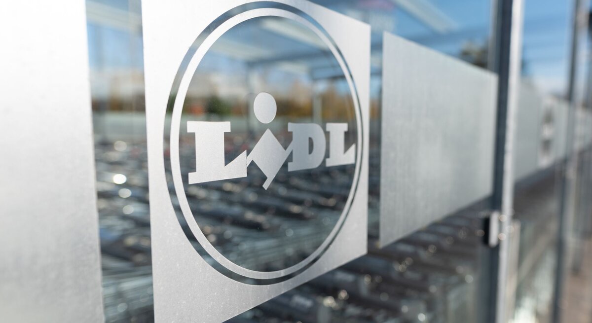 Das Foto zeigt das Logo von Lidl auf einer Glasscheibe, wahrscheinlich am Eingang eines Lidl-Marktes. Das Logo ist mattiert auf der durchsichtigen Fläche, und im Hintergrund sind verschwommen Einkaufswagen zu erkennen, typisch für den Außenbereich eines Supermarktes. Die Aufnahme spielt mit der Tiefenschärfe und fokussiert das Logo, während die Umgebung unscharf bleibt.