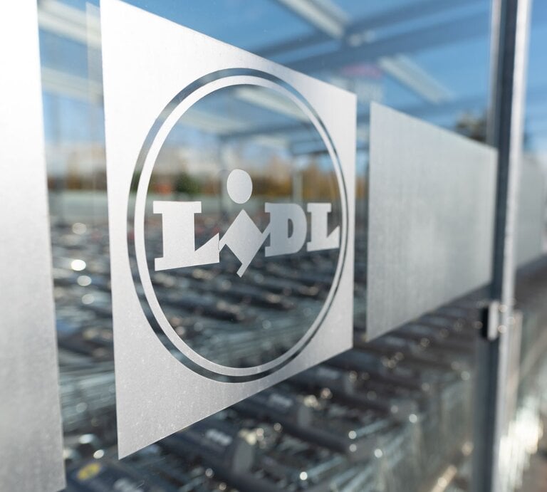 Das Foto zeigt das Logo von Lidl auf einer Glasscheibe, wahrscheinlich am Eingang eines Lidl-Marktes. Das Logo ist mattiert auf der durchsichtigen Fläche, und im Hintergrund sind verschwommen Einkaufswagen zu erkennen, typisch für den Außenbereich eines Supermarktes. Die Aufnahme spielt mit der Tiefenschärfe und fokussiert das Logo, während die Umgebung unscharf bleibt. | © Adobe Stock/ Proxima Studio
