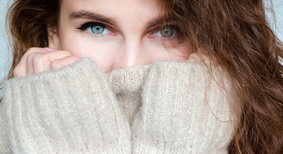 Junge Frau mit lockigem braunem Haar schaut mit hellblauen Augen über den Kragen ihres beigen Strickpullovers, nahes Porträt.