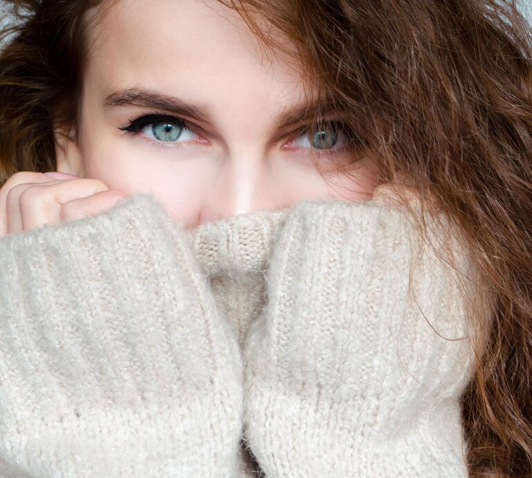 Junge Frau mit lockigem braunem Haar schaut mit hellblauen Augen über den Kragen ihres beigen Strickpullovers, nahes Porträt. | © Adobe Stock/ Svetlana