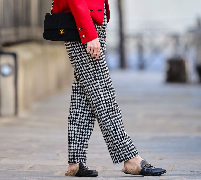 Zu sehen sind lange Beine, die mit einer Hahnentritthose bekleidet sind . Dazu trägt die Frau eine rote Jacke und eine schwarze Tasche.  | © GettyImages/Edward Berthelot / Kontributor