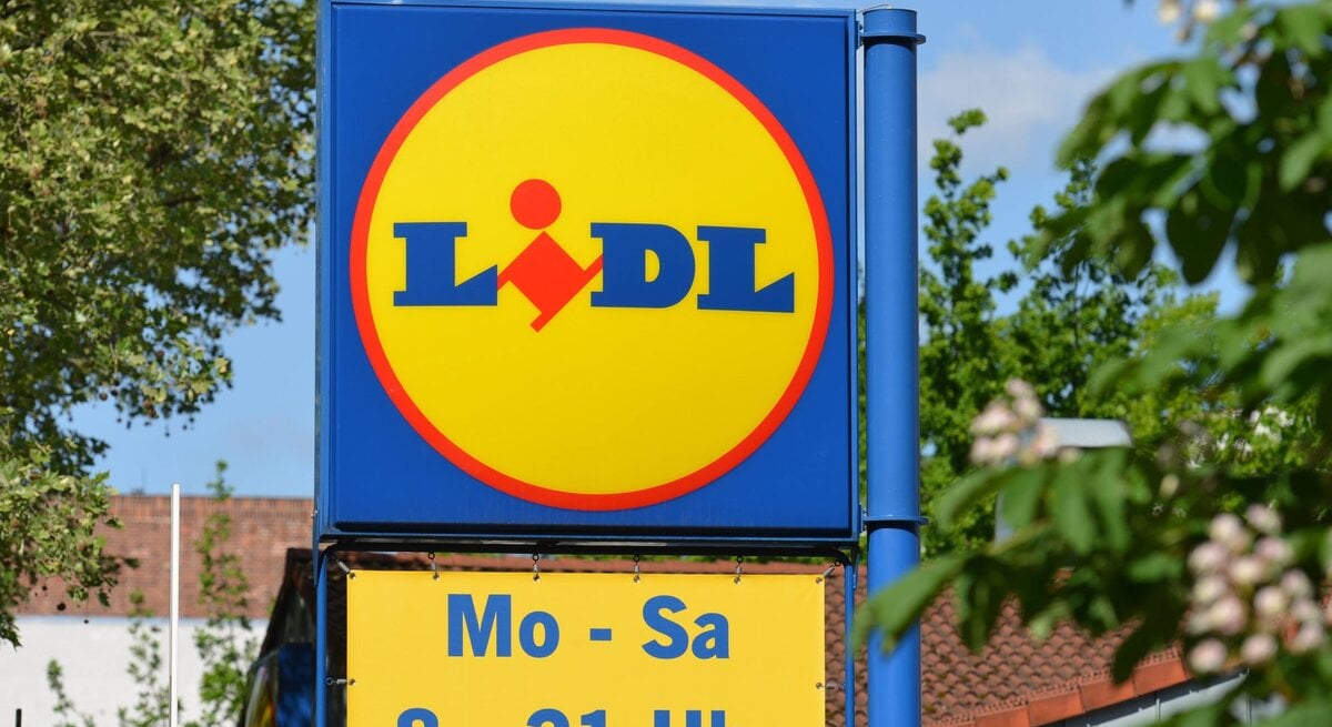 Werbeschild von Lidl mit gelbem Logo und Öffnungszeiten, im Hintergrund grünes Laub und blauer Himmel.