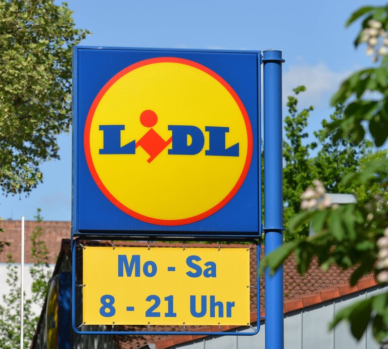 Werbeschild von Lidl mit gelbem Logo und Öffnungszeiten, im Hintergrund grünes Laub und blauer Himmel. | © IMAGO / Schöning