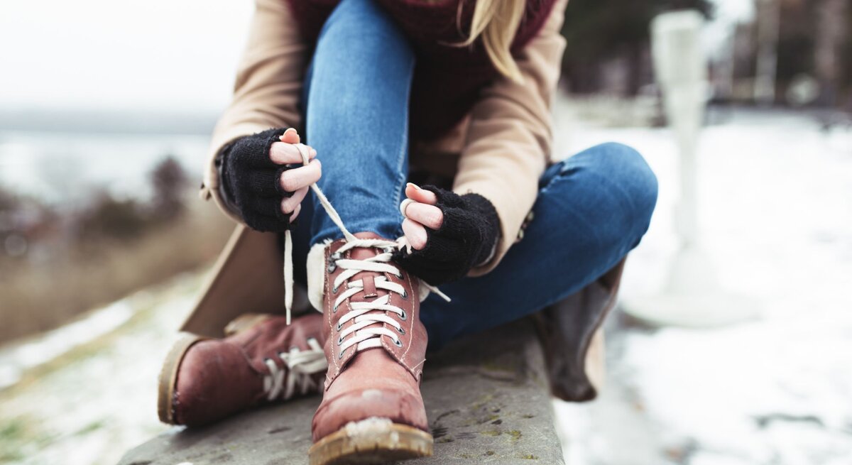 Eine Frau in Jeans und beigem Mantel schnürt braune Winterstiefel, sitzend auf einer Steinbank in verschneiter Landschaft.