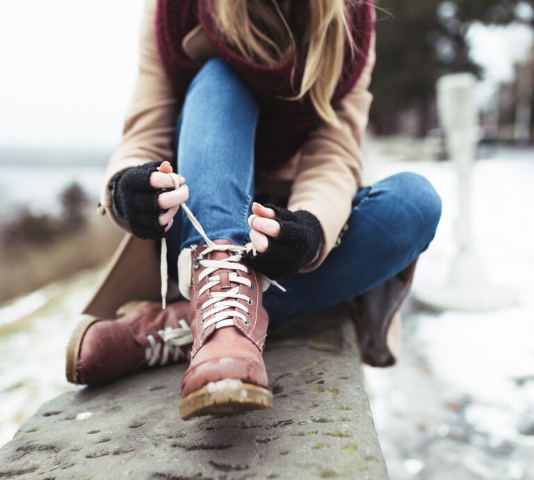 Eine Frau in Jeans und beigem Mantel schnürt braune Winterstiefel, sitzend auf einer Steinbank in verschneiter Landschaft. | © Adobe Stock/ hedgehog94