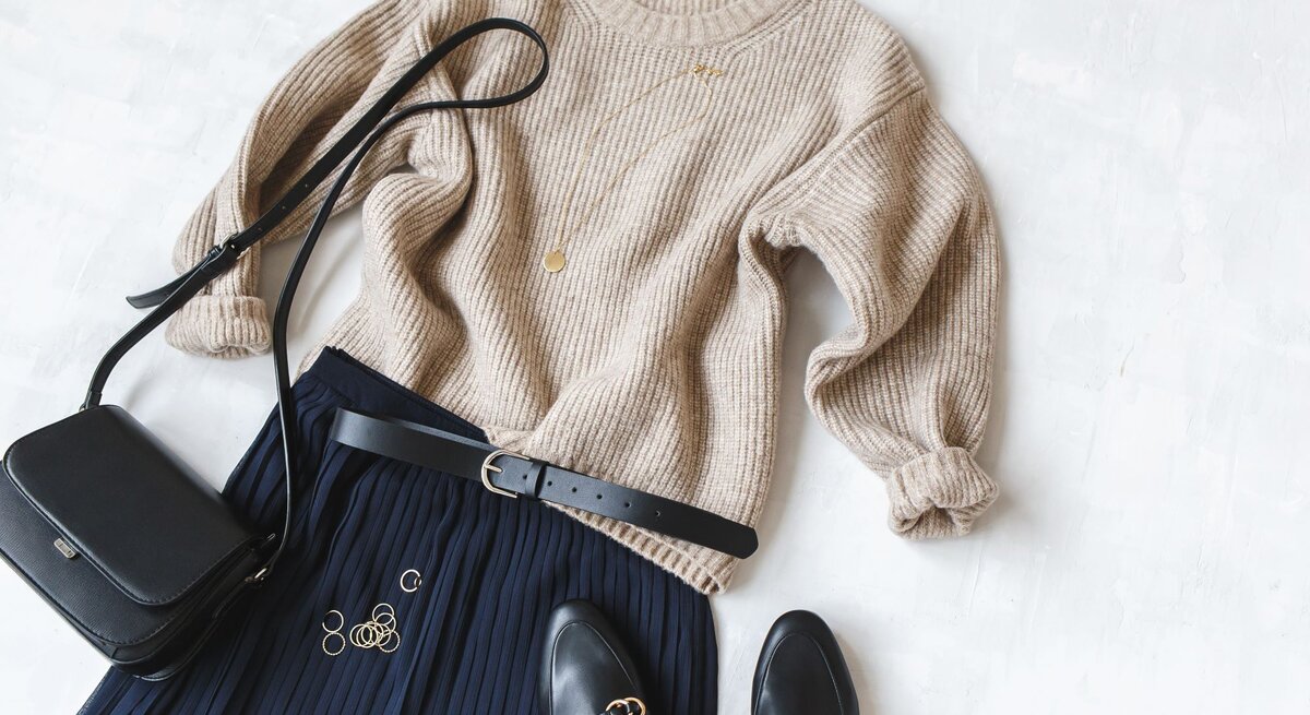Elegantes Outfit aus beigem Pullover, dunkelblauem Strickrock und Accessoires wie goldenen Ringen, Tasche und Loafers auf weißer Fläche.