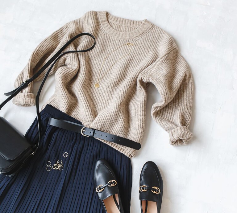 Elegantes Outfit aus beigem Pullover, dunkelblauem Strickrock und Accessoires wie goldenen Ringen, Tasche und Loafers auf weißer Fläche. | © AdobeStock/ NesolenayaAleksandra