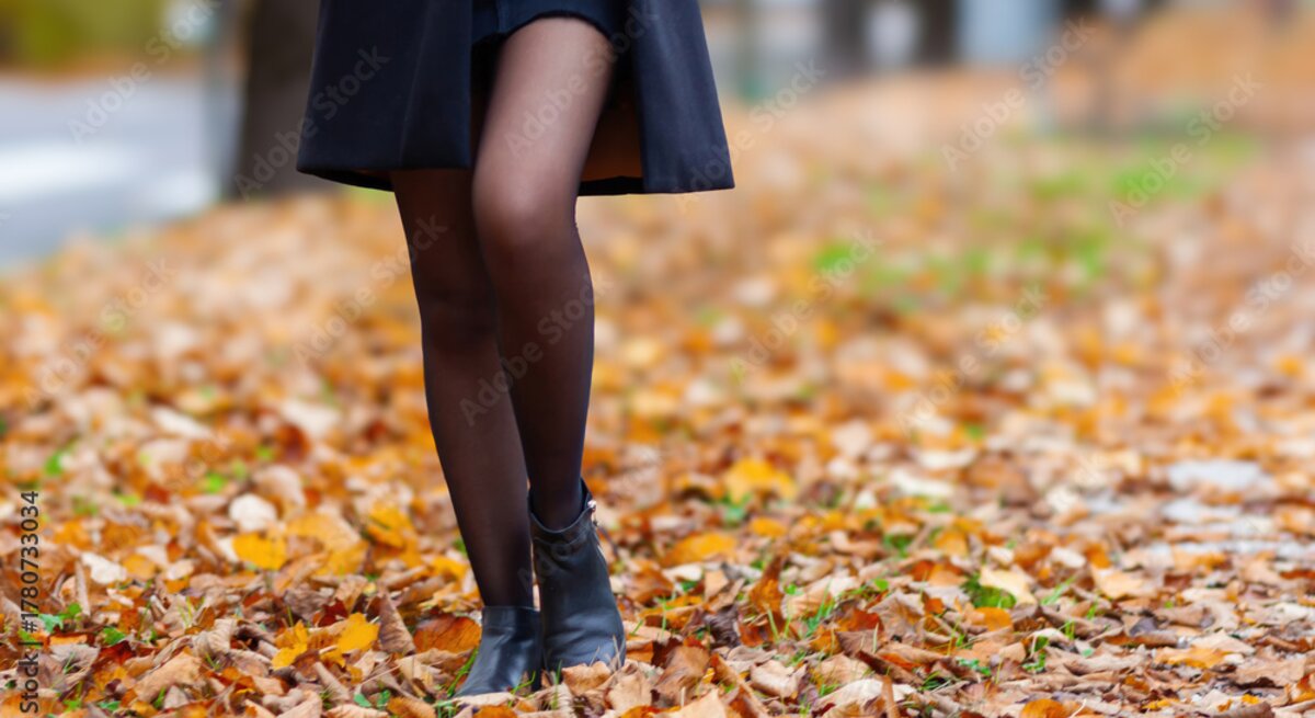 Beine einer Frau in schwarzem Mantel, kurzem Rock und Stiefeletten, gehend über eine von Herbstlaub bedeckte Straße.