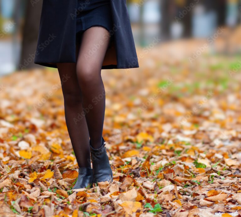 Beine einer Frau in schwarzem Mantel, kurzem Rock und Stiefeletten, gehend über eine von Herbstlaub bedeckte Straße. | © Adobe Stock/ Antonio Gravante