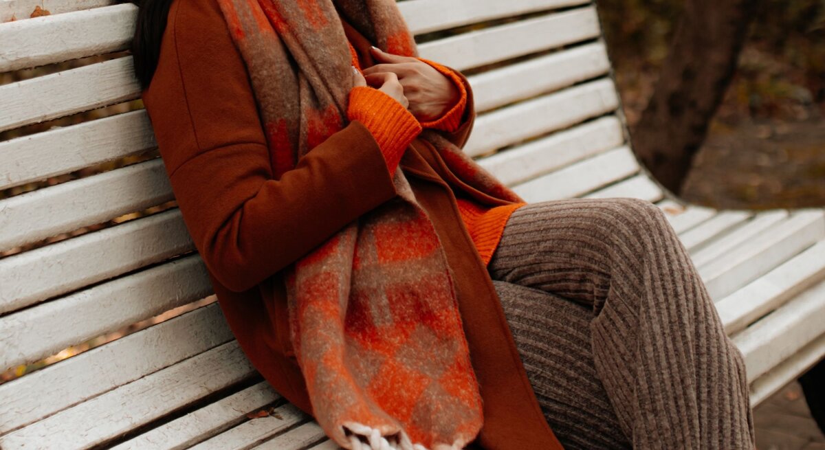 Eine Frau in braunem Mantel, orangefarbenem Schal und Strickhose sitzt entspannt auf einer weißen Parkbank im Herbst.