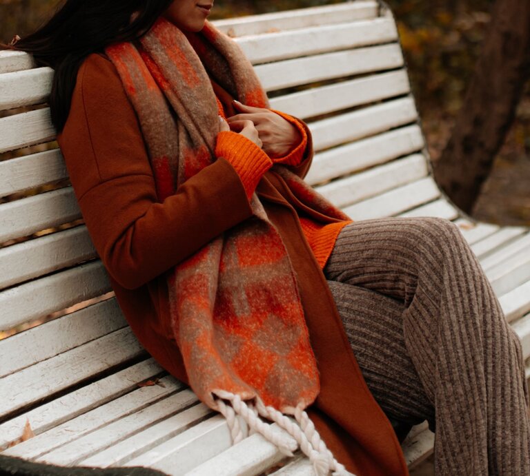 Eine Frau in braunem Mantel, orangefarbenem Schal und Strickhose sitzt entspannt auf einer weißen Parkbank im Herbst. | © Adobe Stock/ asauriet