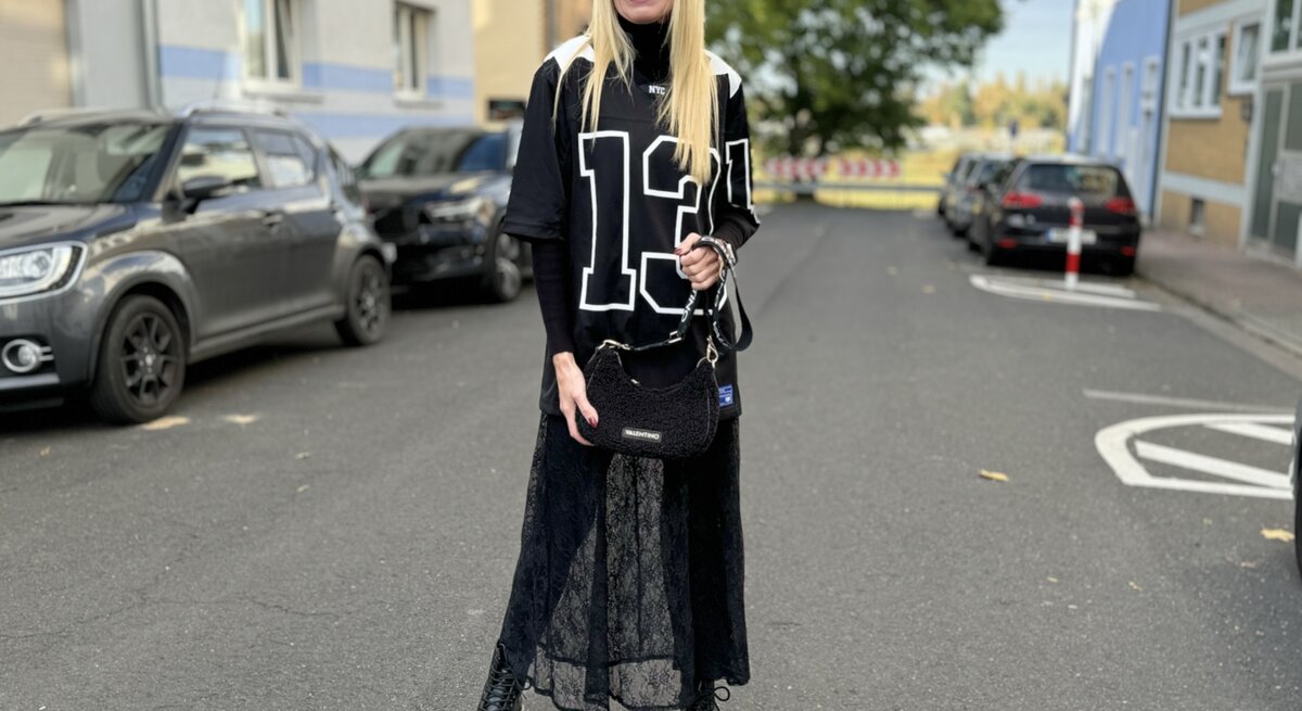 Frau in einem sportlich-modischen Outfit, das an den Blokecore-Trend angelehnt ist. Sie trägt ein übergroßes, schwarz-weißes Jersey mit der Nummer "13", kombiniert mit einem langen Spitzenrock und robusten, schwarzen Stiefeln.