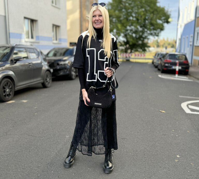 Frau in einem sportlich-modischen Outfit, das an den Blokecore-Trend angelehnt ist. Sie trägt ein übergroßes, schwarz-weißes Jersey mit der Nummer "13", kombiniert mit einem langen Spitzenrock und robusten, schwarzen Stiefeln. | © Renate Zott