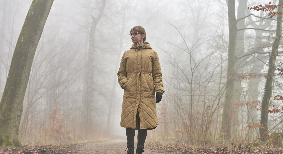 Ältere Frau mit braunem Steppmantel und Handschuhen spaziert nachdenklich auf einem nebligen Waldweg im Herbst.
