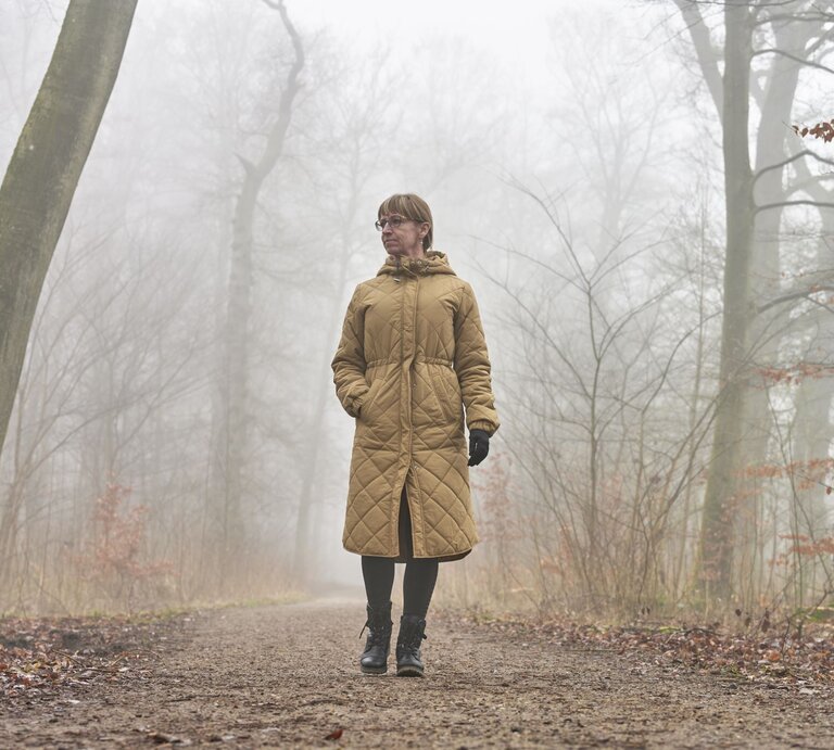 Ältere Frau mit braunem Steppmantel und Handschuhen spaziert nachdenklich auf einem nebligen Waldweg im Herbst. | © Getty Images/ Westersoe