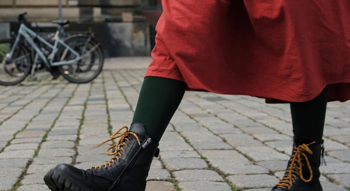 Schwarze klobige Dr Martens Stiefel mit senffarbenen Schnürsenkeln, getragen zu grün-schwarzen Strumpfhosen und einem roten Rock auf Kopfsteinpflaster.