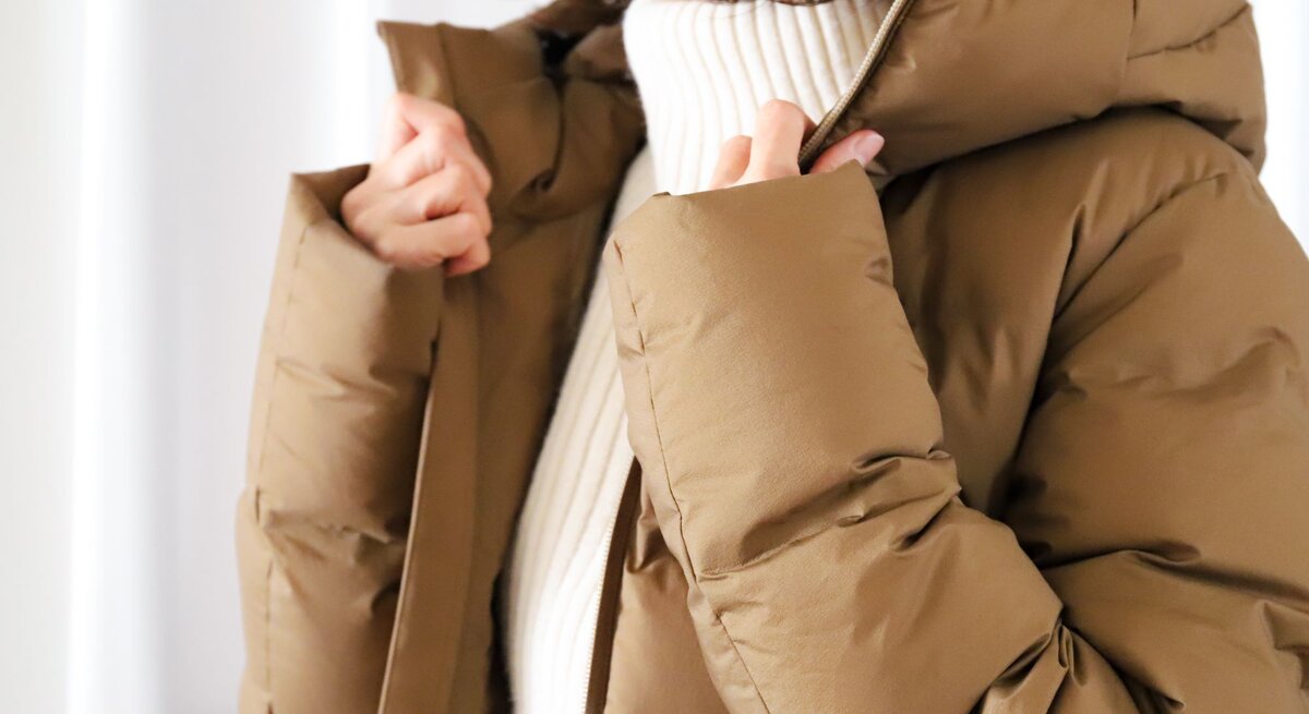 Nahaufnahme einer Person, die eine beige Winterjacke mit Kapuze anzieht, darunter ein weißer, gerippter Pullover.