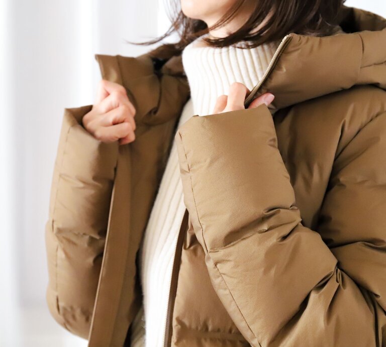 Nahaufnahme einer Person, die eine beige Winterjacke mit Kapuze anzieht, darunter ein weißer, gerippter Pullover. | © Adobe Stock/ webbiz
