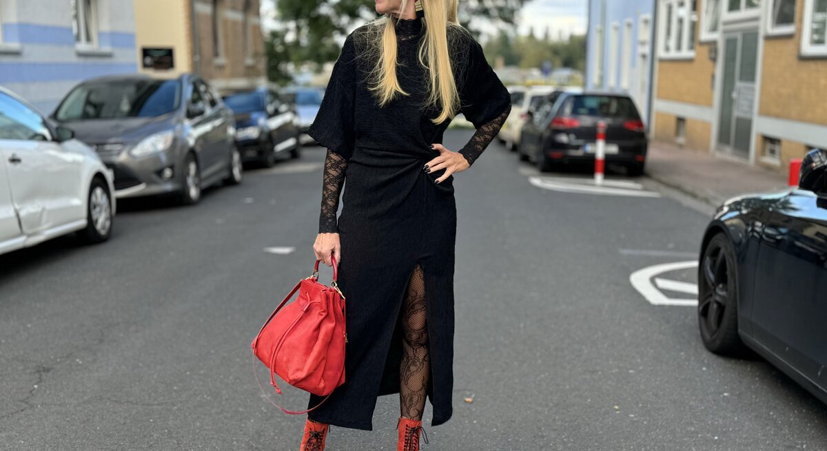 Frau in einem auffälligen Outfit auf der Straße. Sie trägt ein schwarzes Kleid mit Spitzenärmeln und einem hohen Beinschlitz. Dazu kombiniert sie rote Stiefel und eine rote Handtasche, die dem Look farbliche Akzente verleihen. Die Frau trägt ihr blondes Haar offen und eine Sonnenbrille.