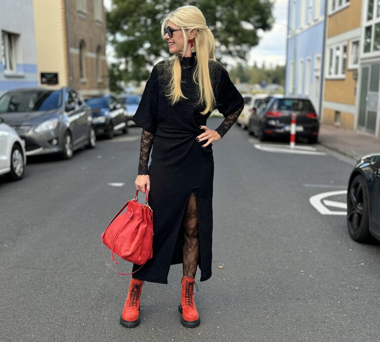 Frau in einem auffälligen Outfit auf der Straße. Sie trägt ein schwarzes Kleid mit Spitzenärmeln und einem hohen Beinschlitz. Dazu kombiniert sie rote Stiefel und eine rote Handtasche, die dem Look farbliche Akzente verleihen. Die Frau trägt ihr blondes Haar offen und eine Sonnenbrille. | © Renate Zott