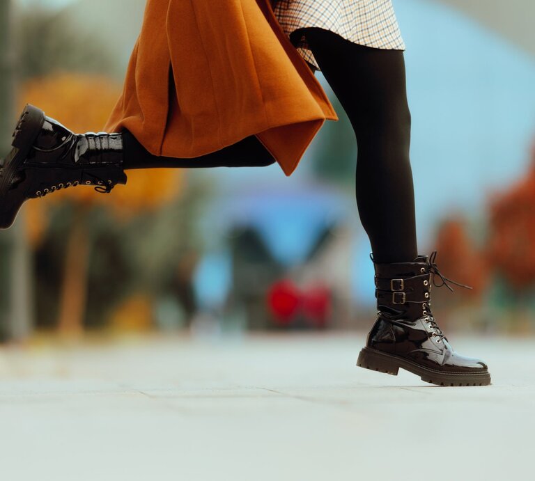 Schwarze, glänzende Stiefel mit dicker Sohle und Schnallen in Bewegung, getragen mit kariertem Rock und braunem Mantel. | © Getty Images/ nicoletaionescu