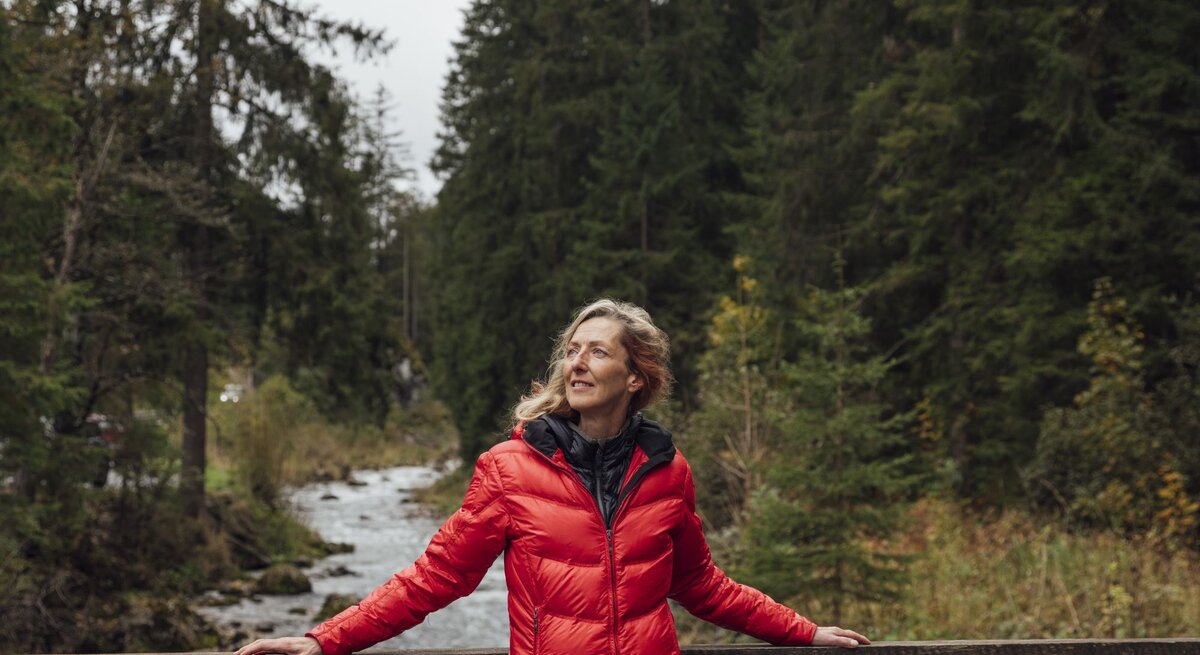 Frau mittleren Alters in roter Jacke lehnt lächelnd an einer Holzbrücke, umgeben von dichtem Wald und Bachlauf.