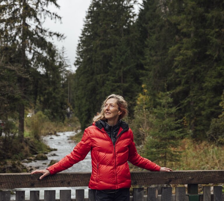 Frau mittleren Alters in roter Jacke lehnt lächelnd an einer Holzbrücke, umgeben von dichtem Wald und Bachlauf. | © Getty Images/ SolStock