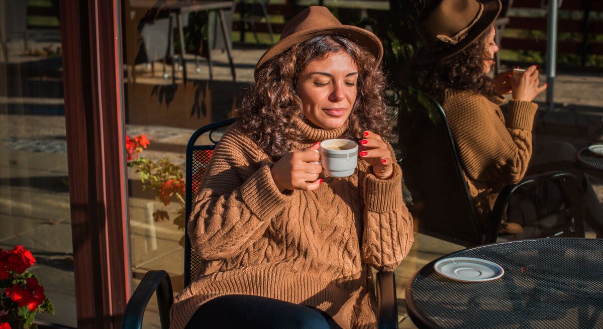 Zu sehen ist eine Frau mittleren Alters, die einen braunen Hut und einen braunen Zopfstrickpullover trägt, während sie Kaffee trinkt