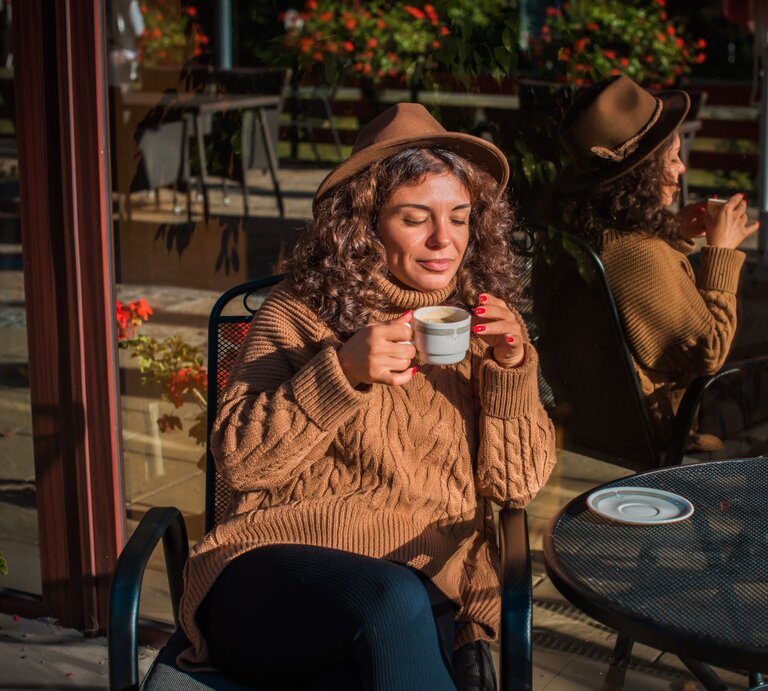 Zu sehen ist eine Frau mittleren Alters, die einen braunen Hut und einen braunen Zopfstrickpullover trägt, während sie Kaffee trinkt | © Shutterstock/T.Den_Team