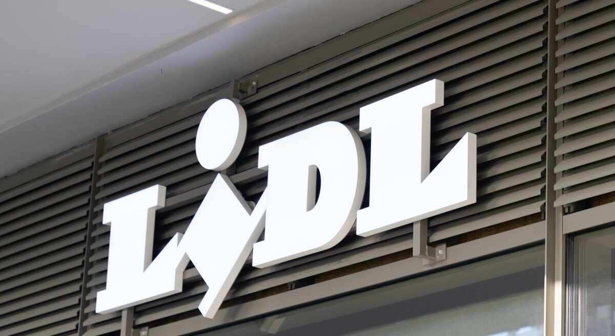 Das Foto zeigt das weiße 3D-Schriftzug-Logo von Lidl, angebracht an einer Ladenfassade über einem Eingang. Das Logo ist vor einem Hintergrund aus horizontalen Lamellen platziert, die typisch für Ladenfronten sind, um Sonnenlicht zu filtern und für Privatsphäre zu sorgen. Die Aufnahme wirkt modern und ist vermutlich tagsüber gemacht worden, wobei das Logo durch seine erhöhte Montage und Farbgebung einen markanten visuellen Akzent setzt.