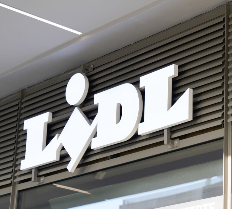 Das Foto zeigt das weiße 3D-Schriftzug-Logo von Lidl, angebracht an einer Ladenfassade über einem Eingang. Das Logo ist vor einem Hintergrund aus horizontalen Lamellen platziert, die typisch für Ladenfronten sind, um Sonnenlicht zu filtern und für Privatsphäre zu sorgen. Die Aufnahme wirkt modern und ist vermutlich tagsüber gemacht worden, wobei das Logo durch seine erhöhte Montage und Farbgebung einen markanten visuellen Akzent setzt. | © Adobe Stock/ OceanProd