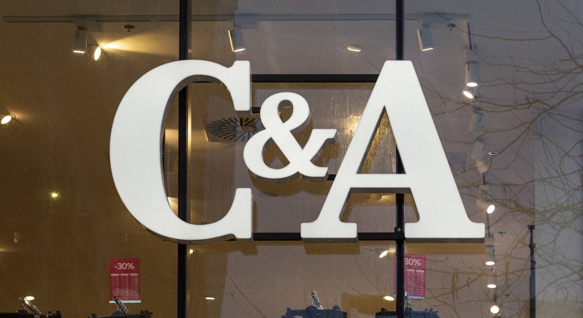  Das Foto zeigt das große, weiße Logo von C&A, das an der Innenseite einer Fensterfront eines Geschäfts angebracht ist. Man kann durch das Fenster in den Laden blicken und einige Kleidungsstücke und Verkaufsschilder mit Rabattangaben sehen, die im Inneren ausgestellt sind. Die Reflexion der Bäume auf der Fensterscheibe lässt darauf schließen, dass das Foto von außen bei Tageslicht aufgenommen wurde.