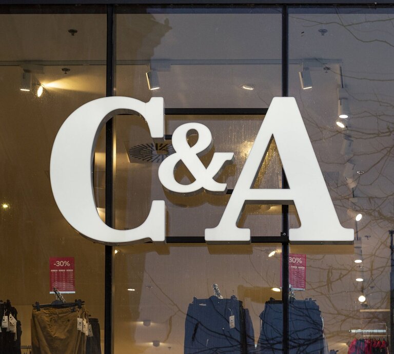  Das Foto zeigt das große, weiße Logo von C&A, das an der Innenseite einer Fensterfront eines Geschäfts angebracht ist. Man kann durch das Fenster in den Laden blicken und einige Kleidungsstücke und Verkaufsschilder mit Rabattangaben sehen, die im Inneren ausgestellt sind. Die Reflexion der Bäume auf der Fensterscheibe lässt darauf schließen, dass das Foto von außen bei Tageslicht aufgenommen wurde. | © IMAGO / CHROMORANGE