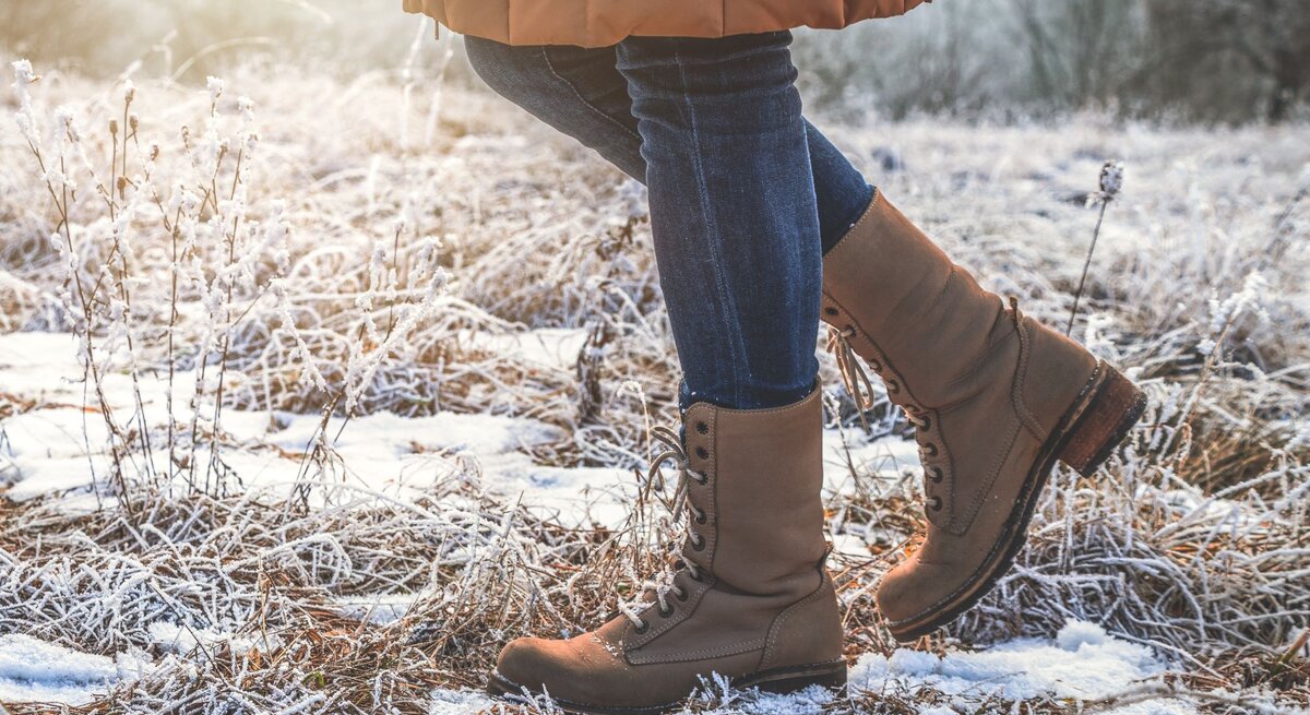 Detailaufnahme von Stiefeln und Jeans, die durch gefrorenes Gras und Schneeflecken in goldenem Morgenlicht schreiten.