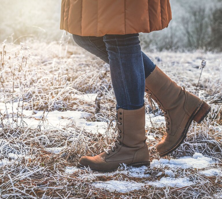 Detailaufnahme von Stiefeln und Jeans, die durch gefrorenes Gras und Schneeflecken in goldenem Morgenlicht schreiten. | © Adobe Stock/ encierro