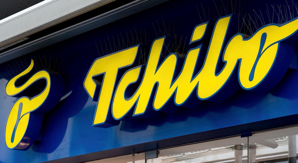 Gelbes Tchibo-Logo auf dunkelblauem Ladenschild, darunter eine Glasfassade mit Spiegelungen von Straßenszenen.
