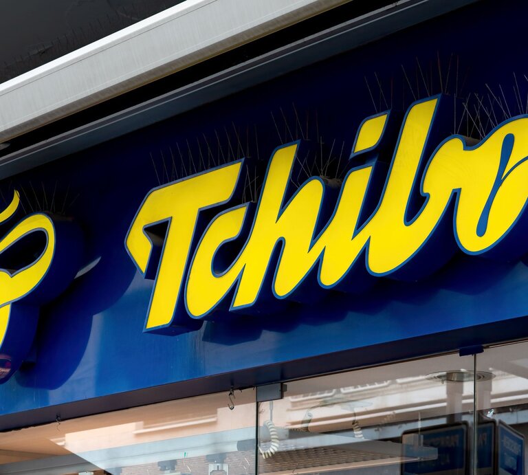 Gelbes Tchibo-Logo auf dunkelblauem Ladenschild, darunter eine Glasfassade mit Spiegelungen von Straßenszenen. | © Adobe Stock/ EKH-Pictures