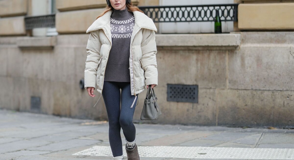 Frau mit beigem Wintermantel, grauem Pullover mit Schneeflockenmuster und Thermoleggings läuft konzentriert über einen Gehweg.