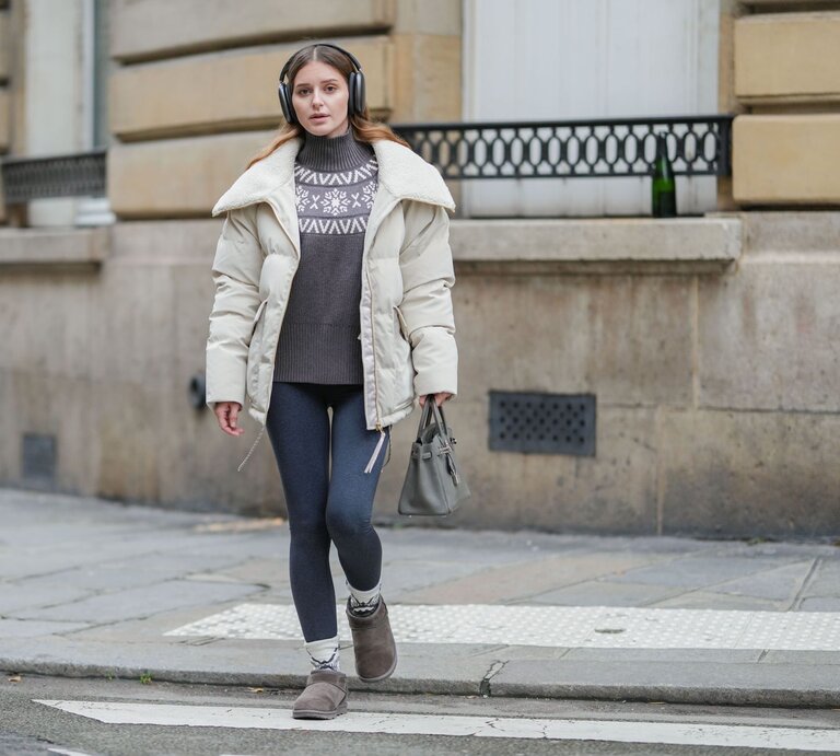 Frau mit beigem Wintermantel, grauem Pullover mit Schneeflockenmuster und Thermoleggings läuft konzentriert über einen Gehweg. | © Edward Berthelot/Getty Images