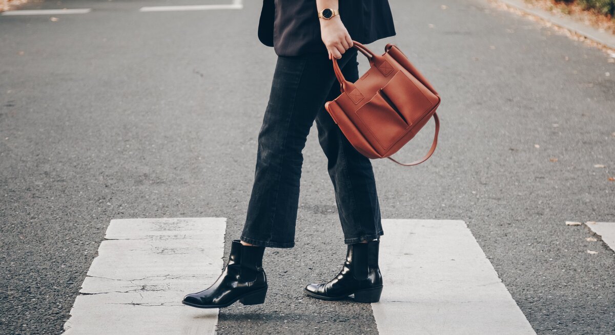 Eine Person trägt schwarze Boots, dunkle Jeans und hält eine braune Tasche, während sie einen Zebrastreifen überquert.