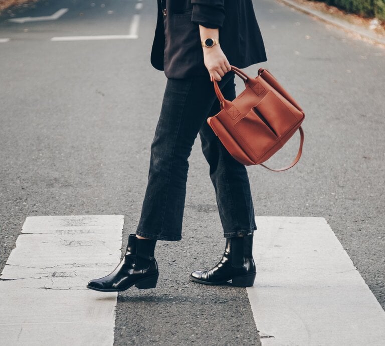 Eine Person trägt schwarze Boots, dunkle Jeans und hält eine braune Tasche, während sie einen Zebrastreifen überquert. | © Adobe Stock/ mlasaimages
