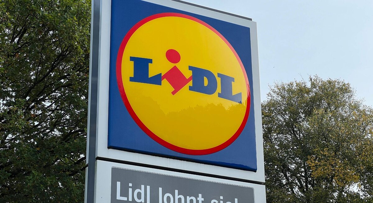 Auf dem Bild ist ein Werbeschild von Lidl zu sehen. Im oberen Teil des Schildes ist das typische Lidl-Logo mit gelbem Hintergrund und dem roten Lidl-Schriftzug. Unterhalb des Logos befindet sich der Slogan „Lidl lohnt sich.“ im Weiß auf einem blauen Hintergrund. Im Hintergrund des Schildes sind Bäume mit grünem Laub zu erkennen, was auf eine Aufnahme bei Tageslicht hindeutet.