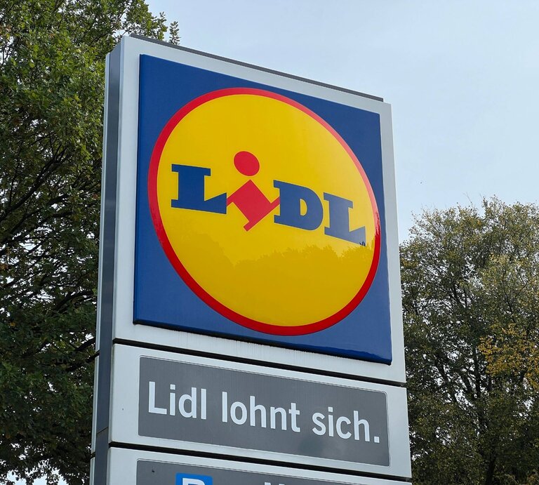 Auf dem Bild ist ein Werbeschild von Lidl zu sehen. Im oberen Teil des Schildes ist das typische Lidl-Logo mit gelbem Hintergrund und dem roten Lidl-Schriftzug. Unterhalb des Logos befindet sich der Slogan „Lidl lohnt sich.“ im Weiß auf einem blauen Hintergrund. Im Hintergrund des Schildes sind Bäume mit grünem Laub zu erkennen, was auf eine Aufnahme bei Tageslicht hindeutet. | © IMAGO / Lobeca