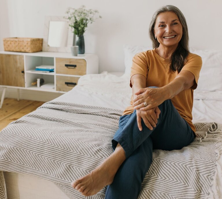 Ältere Frau mit grauen langen Haaren sitzt lächelnd mit Straßenkleidung auf einem Bett in einem hellen Zimmer mit schlichter Dekoration. | © Adobe Stock/ shurkin_son