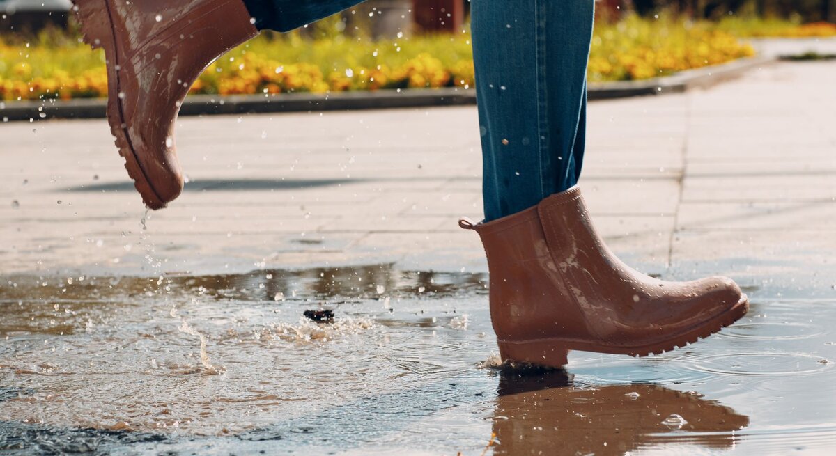 Nahaufnahme von Beinen in Jeans und glänzenden braunen Stiefeln, die schwungvoll durch eine Pfütze auf einem sonnigen Weg treten.