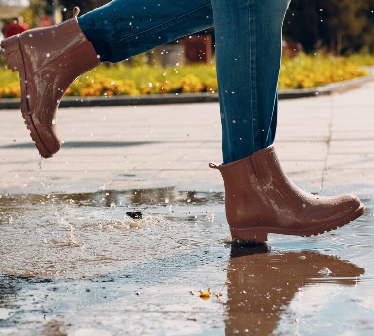 Nahaufnahme von Beinen in Jeans und glänzenden braunen Stiefeln, die schwungvoll durch eine Pfütze auf einem sonnigen Weg treten. | © Adobe Stock/ primipil