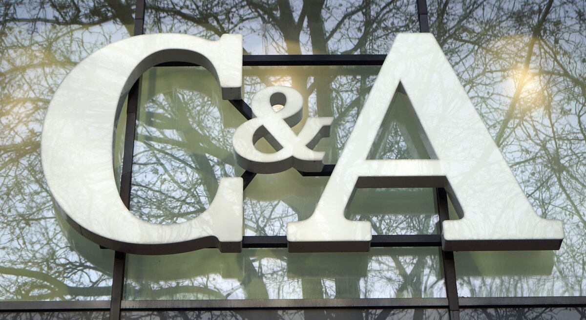 Das Foto zeigt das große weiße Logo "C&A" vor einer Fensterfront, in der sich Bäume spiegeln. Das Logo hebt sich deutlich gegen den durch die Spiegelung erzeugten natürlichen Hintergrund ab. Die Szene wirkt ruhig und die Farben sind harmonisch aufeinander abgestimmt, was einen interessanten visuellen Effekt erzeugt.